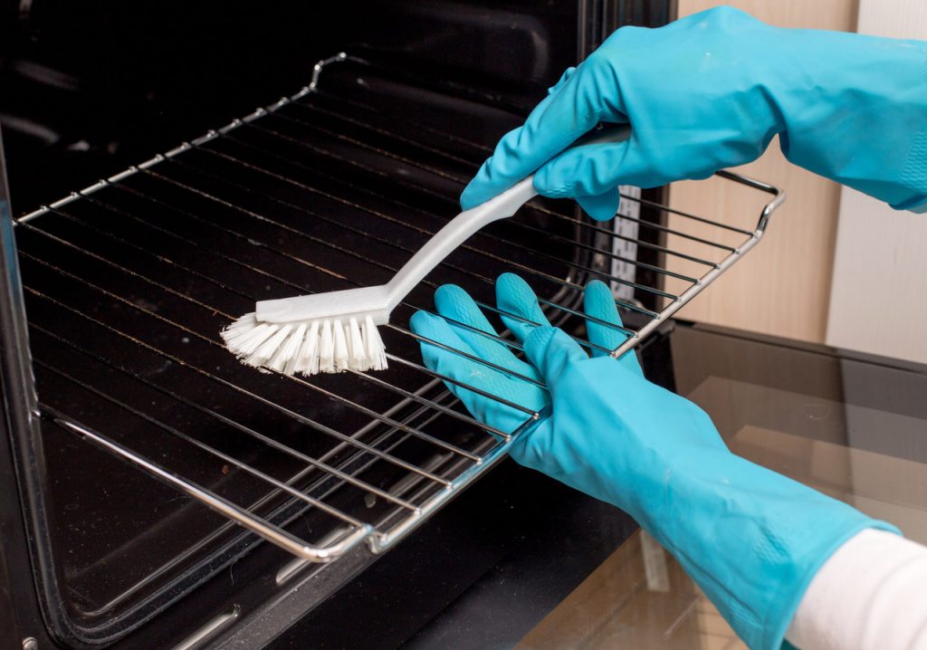ręce w w niebieskich, gumowych rękawiczkach czyszczą ruszt piekarnika za pomocą plastikowej szczotki