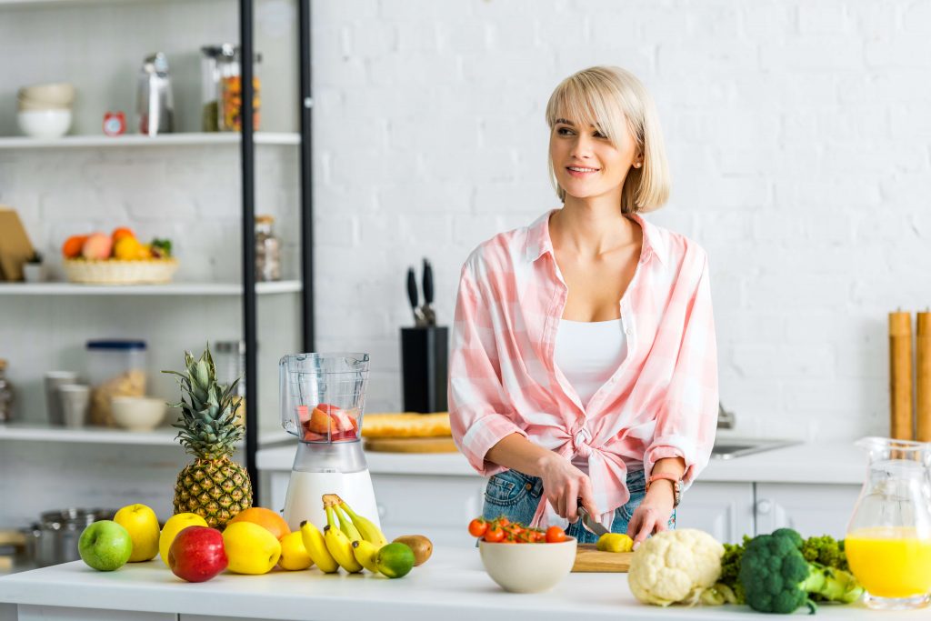 Uśmiechnięta kobieta kroi owoce i warzywa na tle kuchni