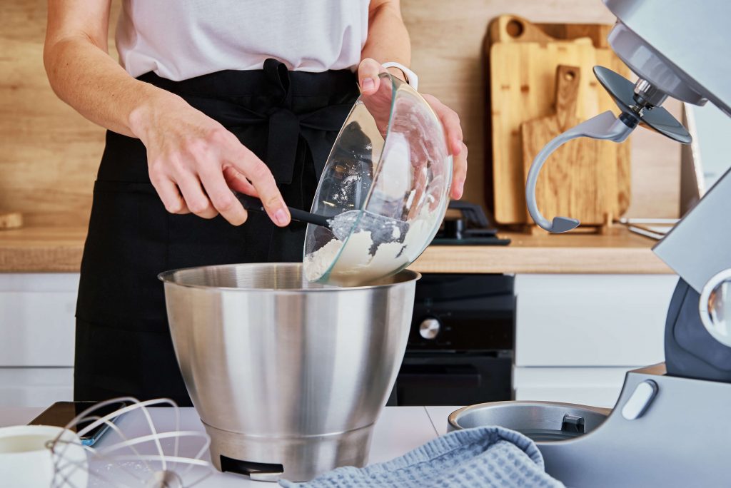kobieta przesypuje mąkę ze szklanej misy do misy robota kuchennego
