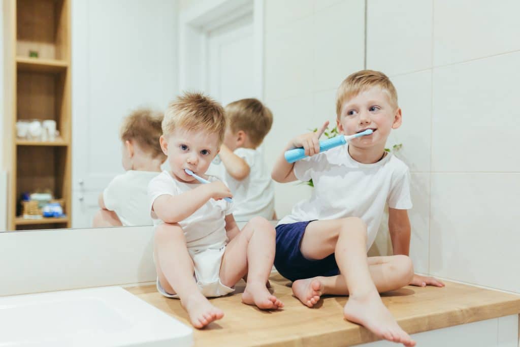 Dwaj chłopcy w wieku ok. 2 i 5 lat, siedzący na blacie w łazience i myjący zęby szczoteczkami dla dzieci