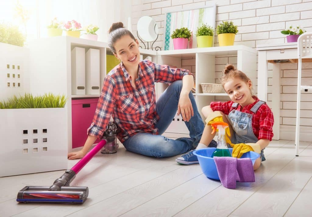 Mama i córka siedzą na podłodze w domu i planują sprzątanie. Obok stoją odkurzacz pionowy i miska ze środkami czystości.