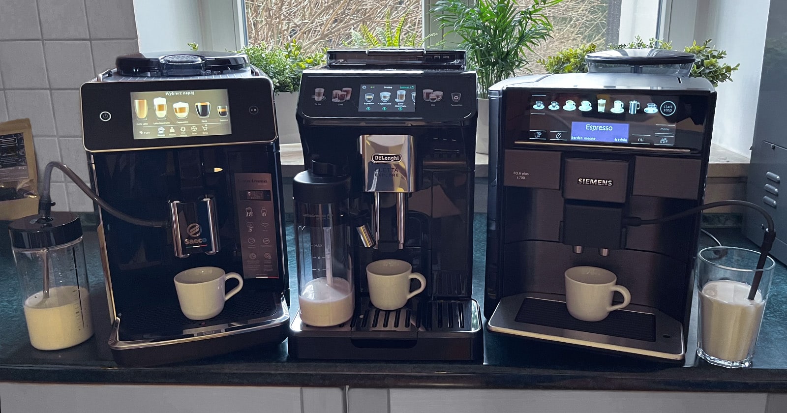 Trzy ekspresy do kawy: Siemens EQ.6 plus s 700 TE657319RW, De’Longhi Eletta Explore ECAM450.55.G i SaecoGranAroma Deluxe SM6680/00 stojące na blacie z białymi kubkami i pełnymi systemami mlecznymi - zdjęcie.