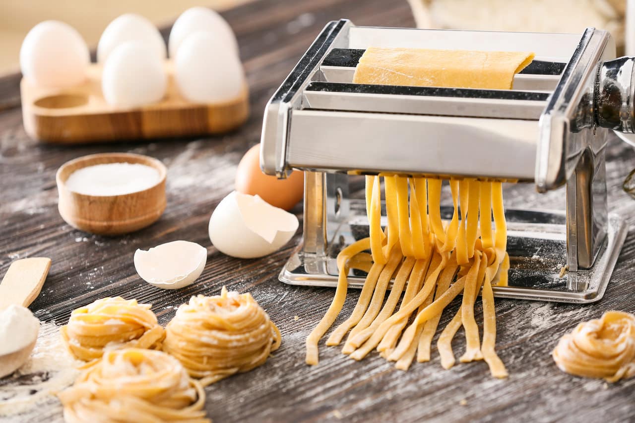 Włoska maszynka do robienia domowego makaronu wstążki, obok jajka, rozsypana mąka i gotowe gniazdka makaronu