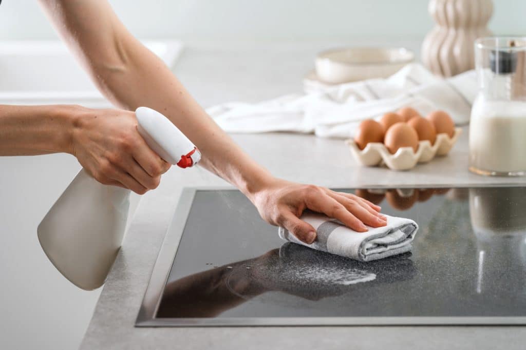 Kobiece dłonie czyszczące płytę indukcyjną ściereczką i detergentem w sprayu