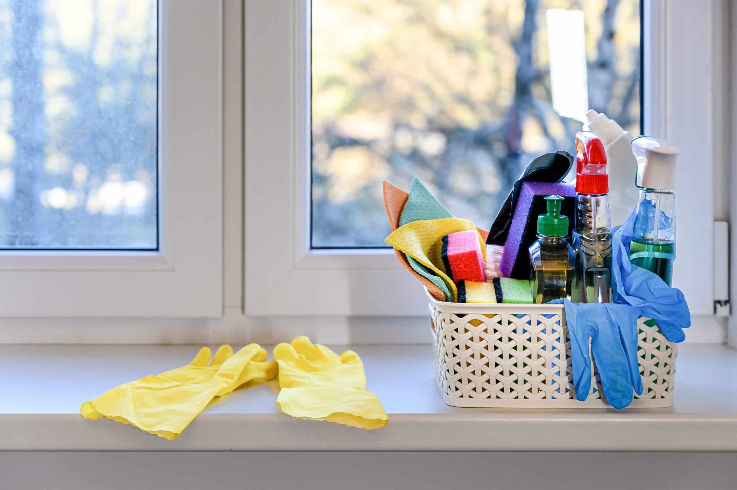 Koszyk ze ściereczkami, gąbkami oraz środkami do mycia okien, przeszkleń i parapetów stojący na okiennym parapecie; w tle brudne okno