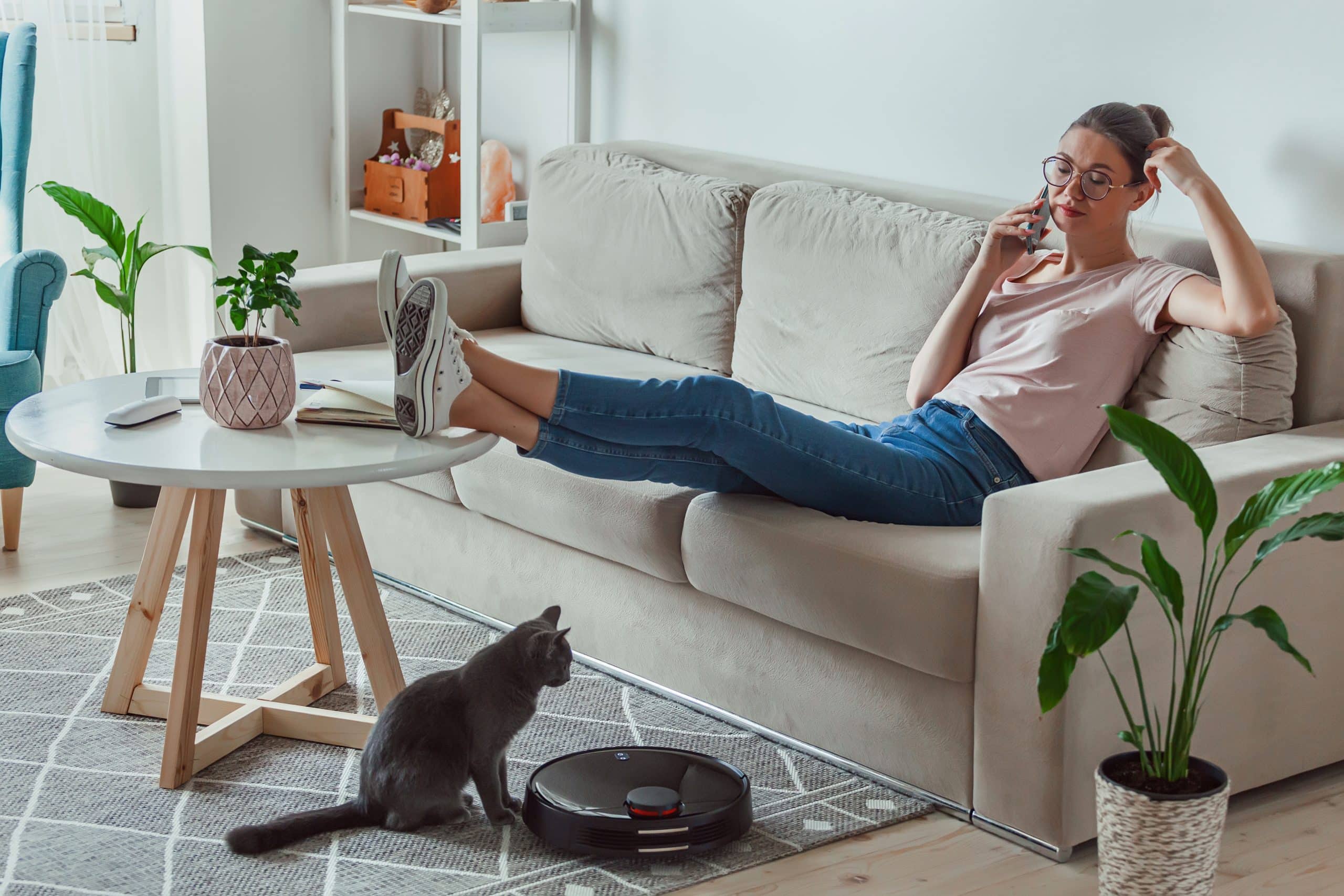 Robot sprzątający na dywanie, obok niego kot i kobieta odpoczywającą na kanapie