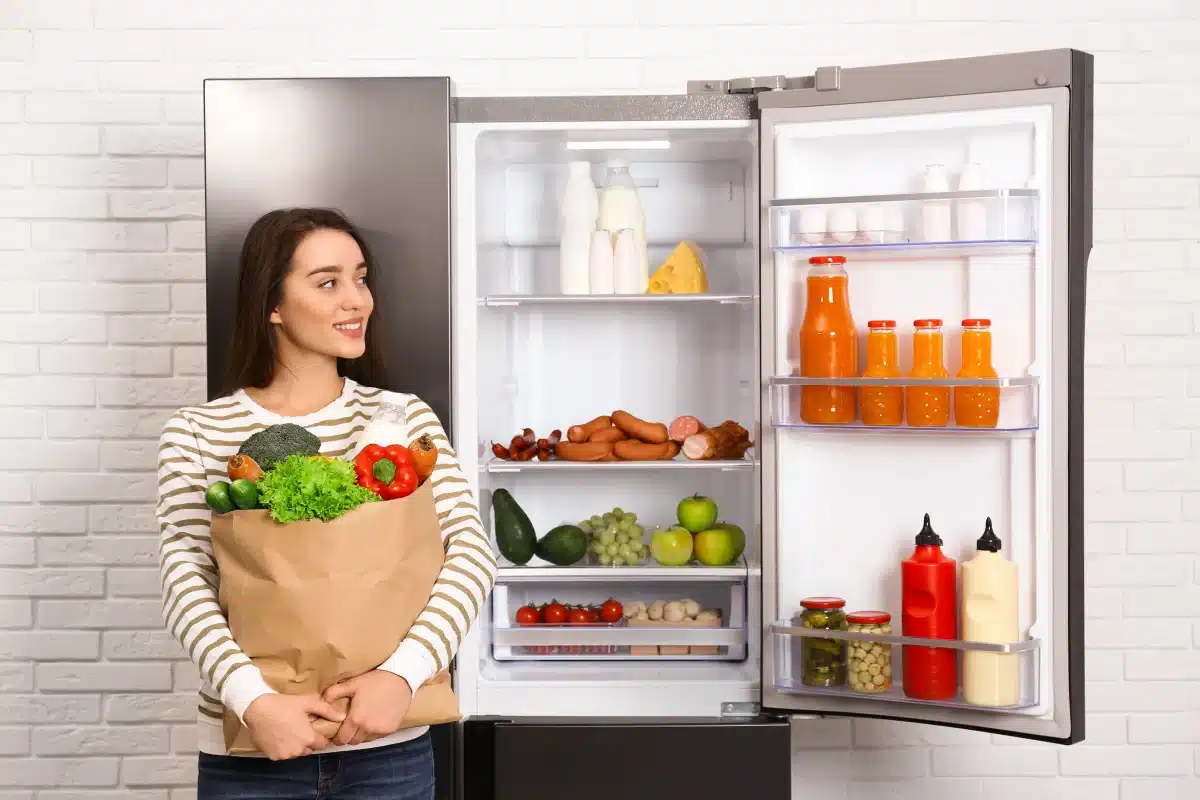 Otwarta lodówka z optymalnie rozmieszczonymi produktami, obok której stoi młoda kobieta z torbą pełną warzyw