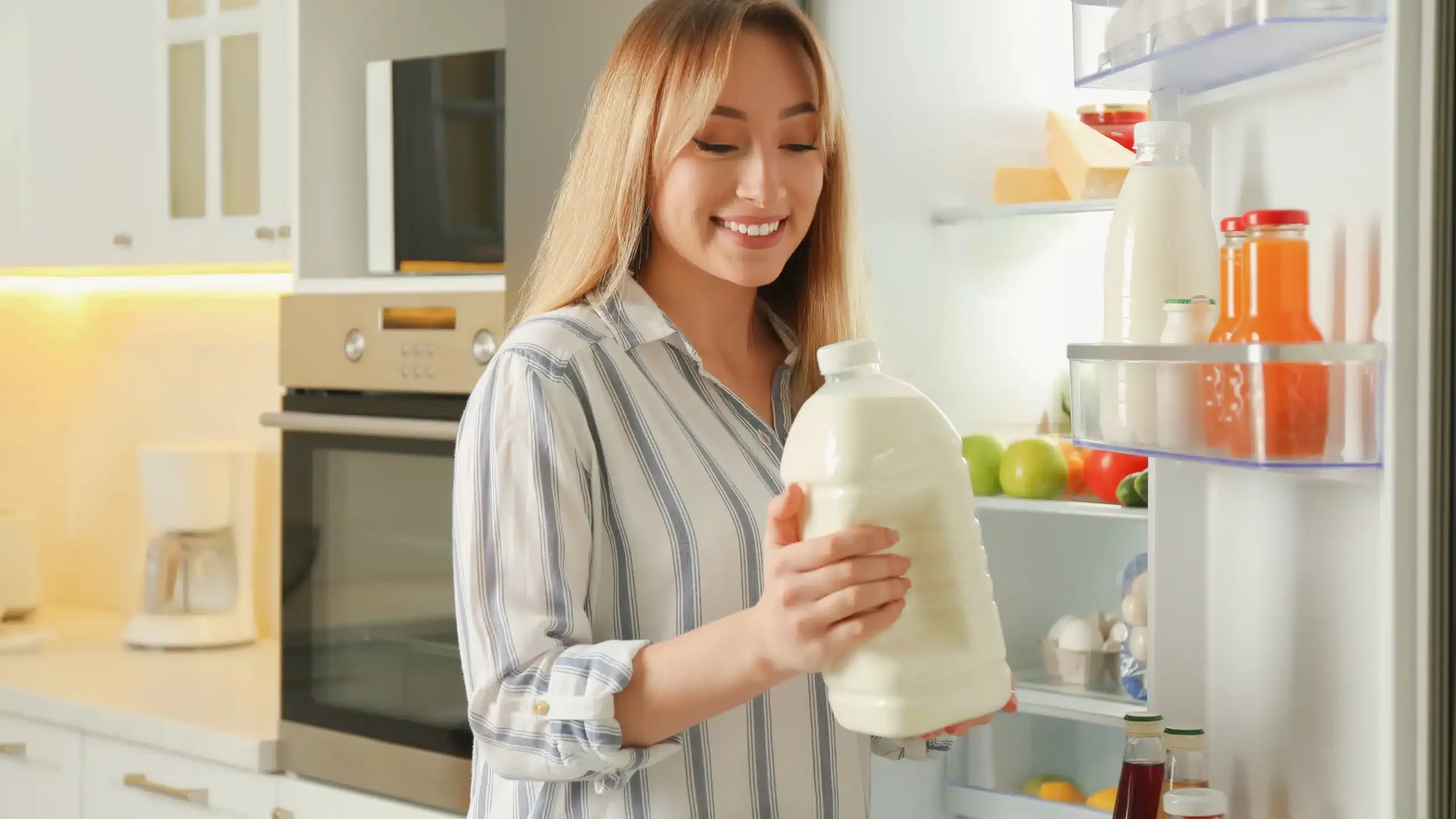 Kobieta trzymająca butelkę mleka stoi przy otwartej lodówce zastanawiając się, jak układać w lodówce nabiał