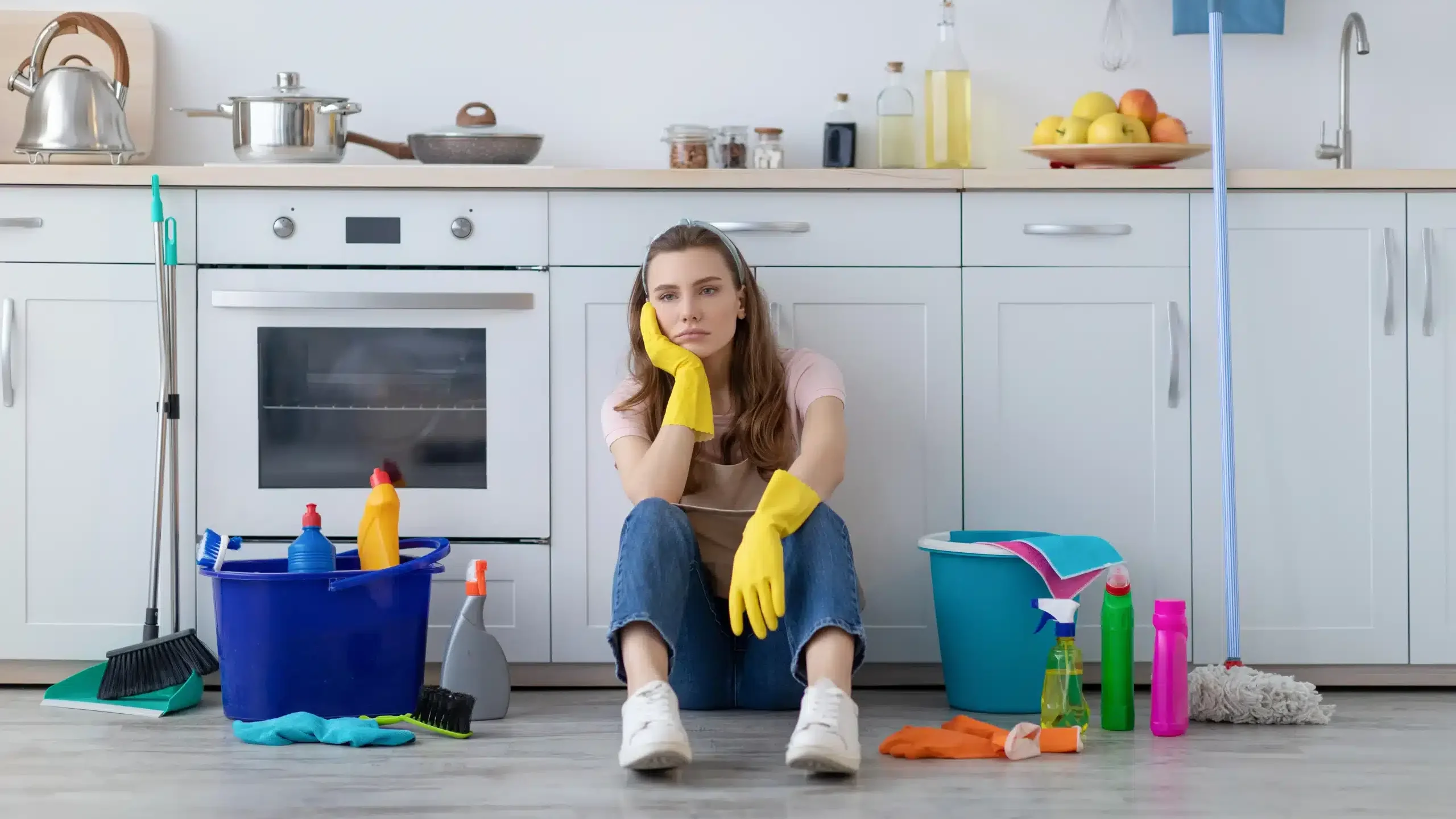 Zniechęcona smugami na podłodze kobieta siedząca przy szafce kuchennej w otoczeniu detergentów i akcesoriów do sprzątania