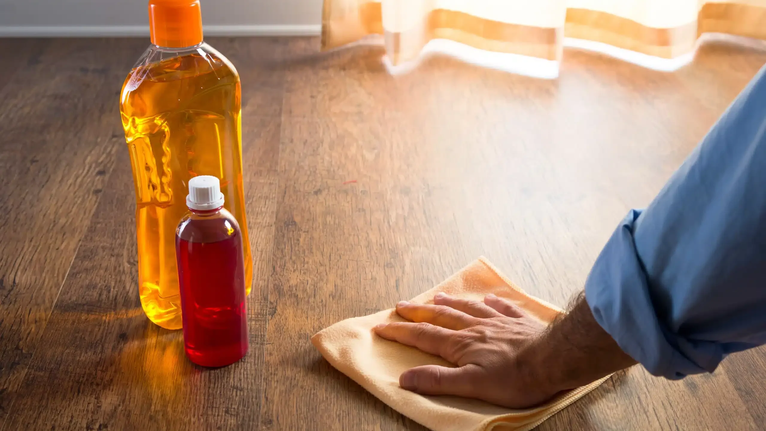 Męska dłoń myje drewnianą podłogę szmatką z mikrofibry; obok 2 butelki preparatów do czyszczenia drewnianej podłogi