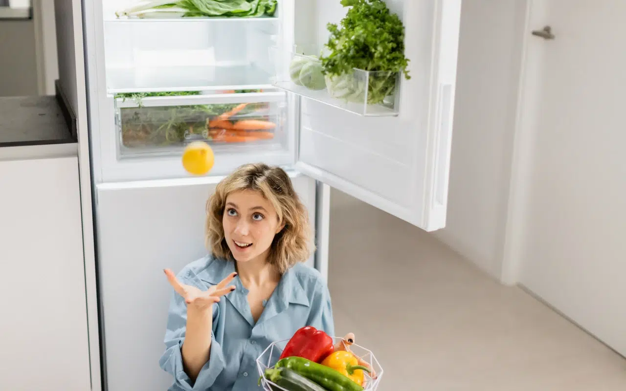 Młoda kobieta trzyma koszyk z warzywami siedząc przy otwartej lodówce dwudrzwiowej z dolnym zamrażalnikiem