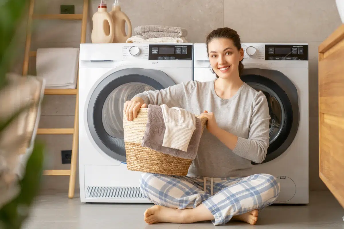 Młoda kobieta z koszem prania siedząca na podłodze w pralni; za nią oddzielne pralka i suszarka