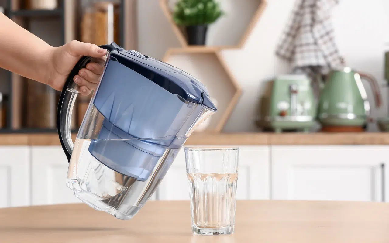 Dłoń trzyma dzbanek do filtrowania wody i nalewa filtrowaną wodę do szklanki stojącej na kuchennym blacie