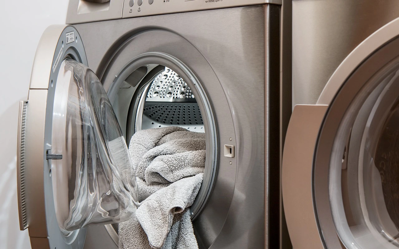 Otwarte drzwiczki srebrnej pralki ładowanej od przodu, z wystającym z bębna szarym ręcznikiem.