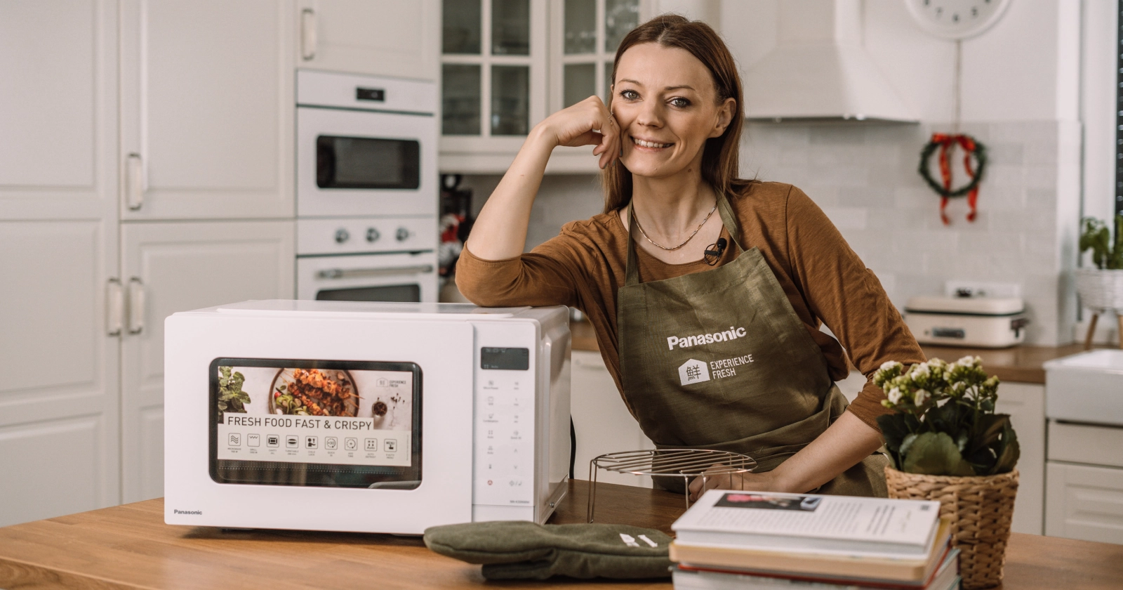 Młoda, radosna kobieta opiera się łokciem o wolnostojącą kuchenkę mikrofalową Panasonic NN-K35NWM