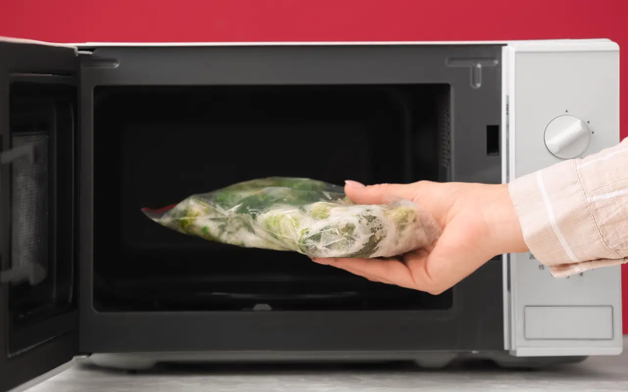 Dłoń wkłada zamrożone warzywa w woreczku do wnętrza mikrofalówki z funkcją rozmrażania