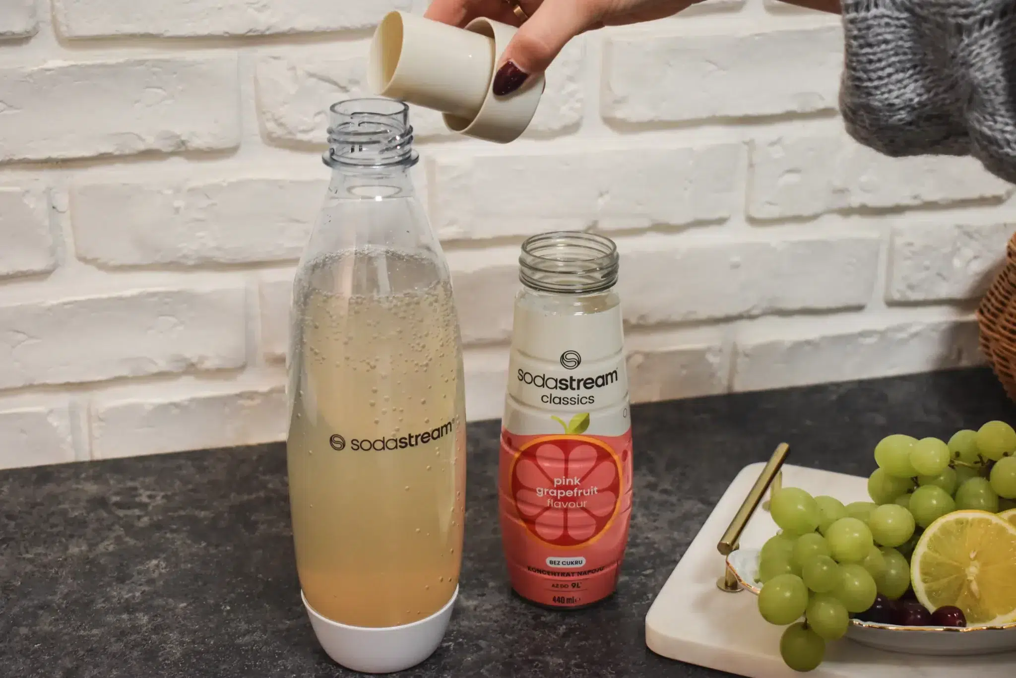 kobieta wlewa syrop SodaStream Różowy Grapefruit do butelki z wodą gazowaną w saturatorze SodaStream